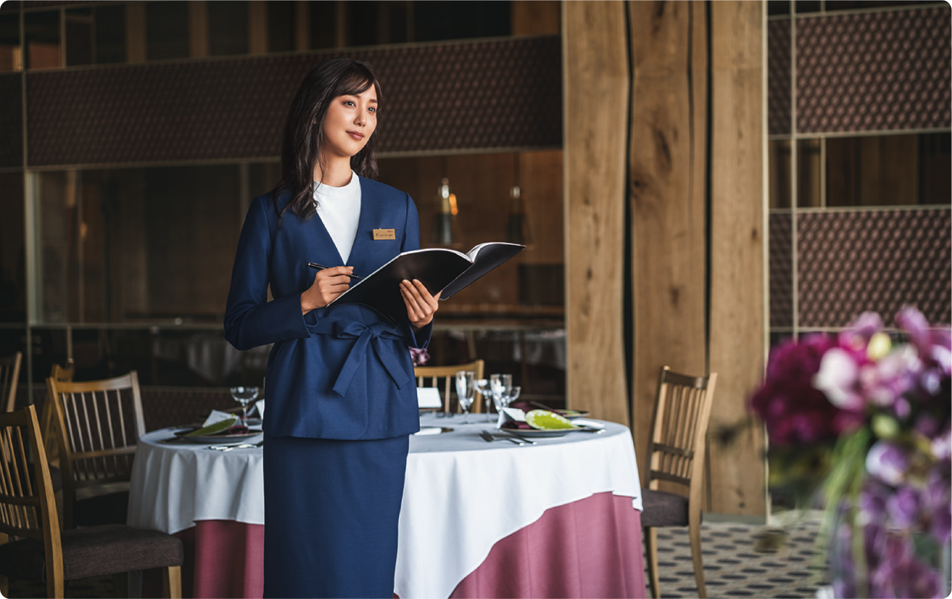 ENJOY Noirのネイビー色の制服を着たウエディングプランナーの女性が、高級感のある会場でイベントの準備を確認する様子